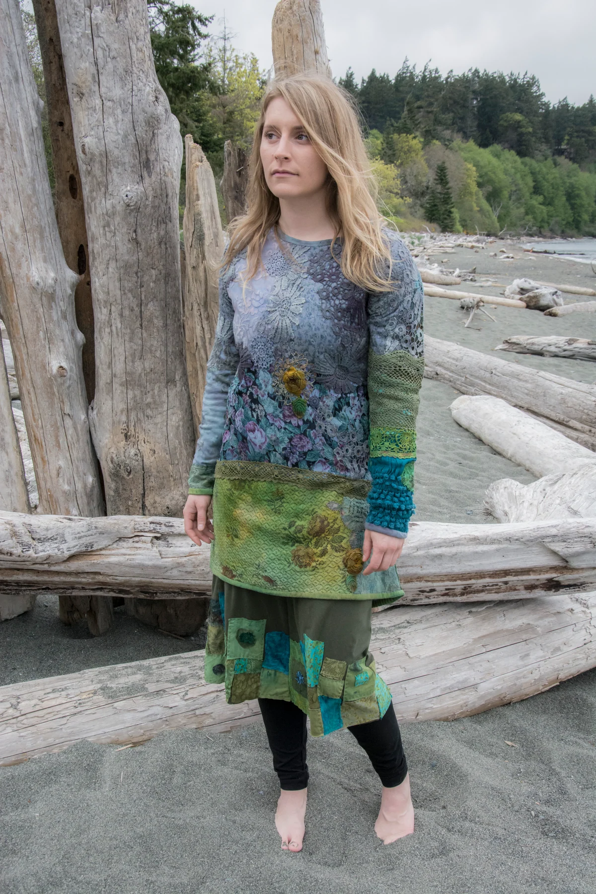 Model wearing a blue-grey lace top and a green patchwork skirt beside driftwood.
