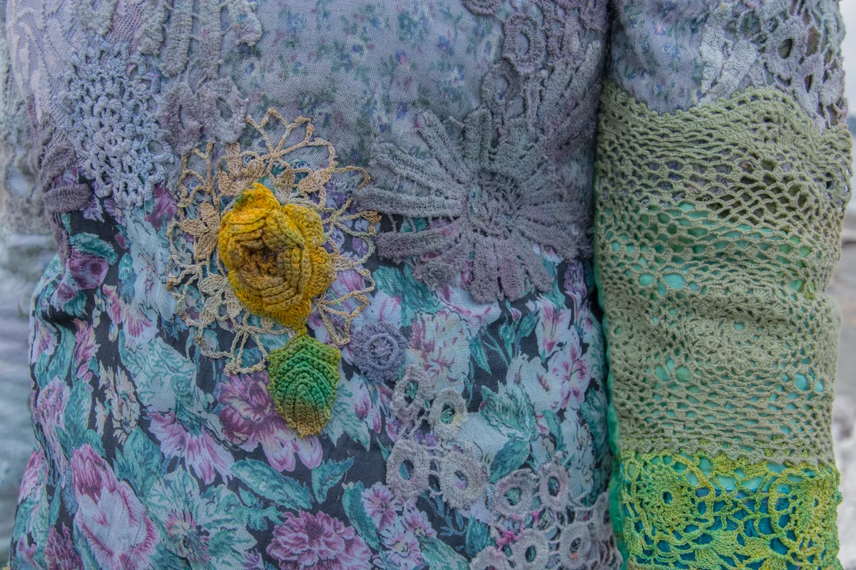Close view of blue-grey lace, floral fabric and a yellow textile embellishment on a garment.