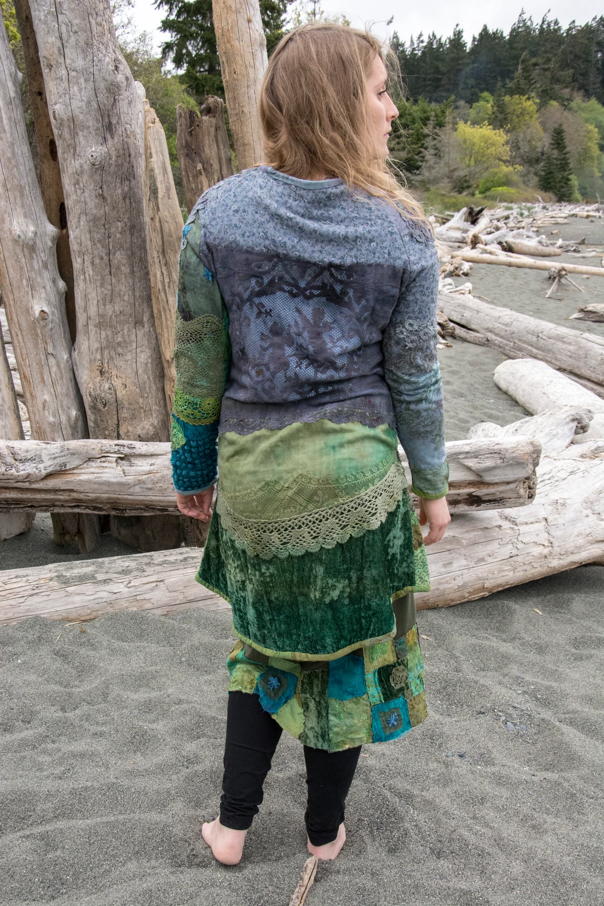 Back view of a model wearing blue-grey and green layered textile garments on the beach.