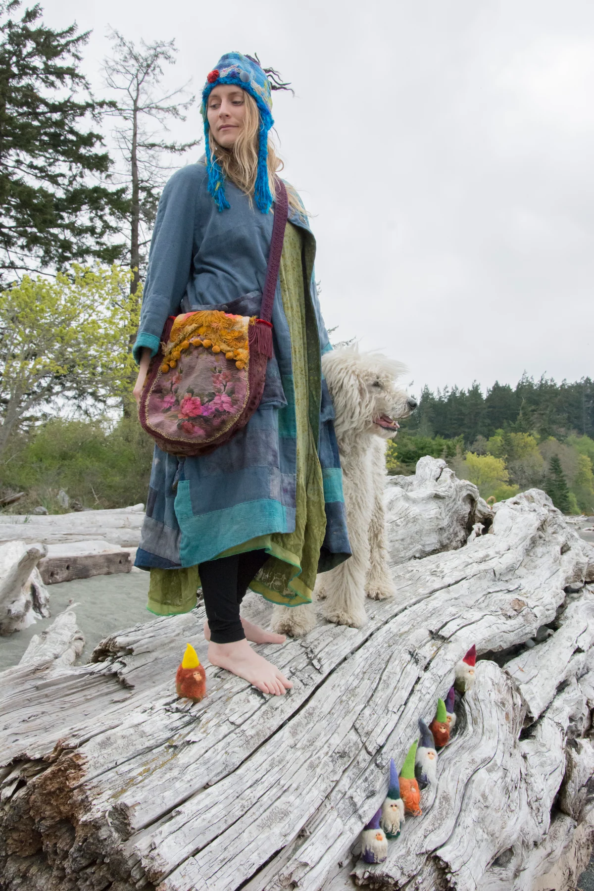 Model wearing a long blue textile outfit, a textured hat and a round embroidered bag on driftwood.