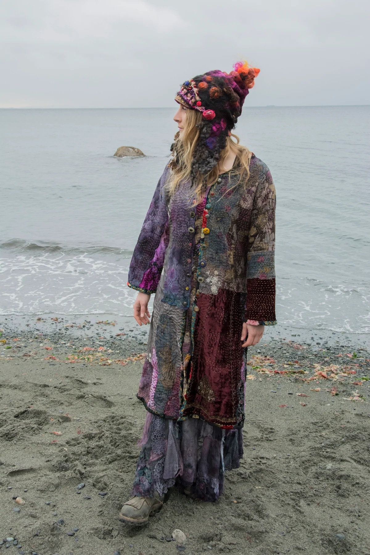 Model wearing a long layered purple and burgundy textile outfit by the sea.