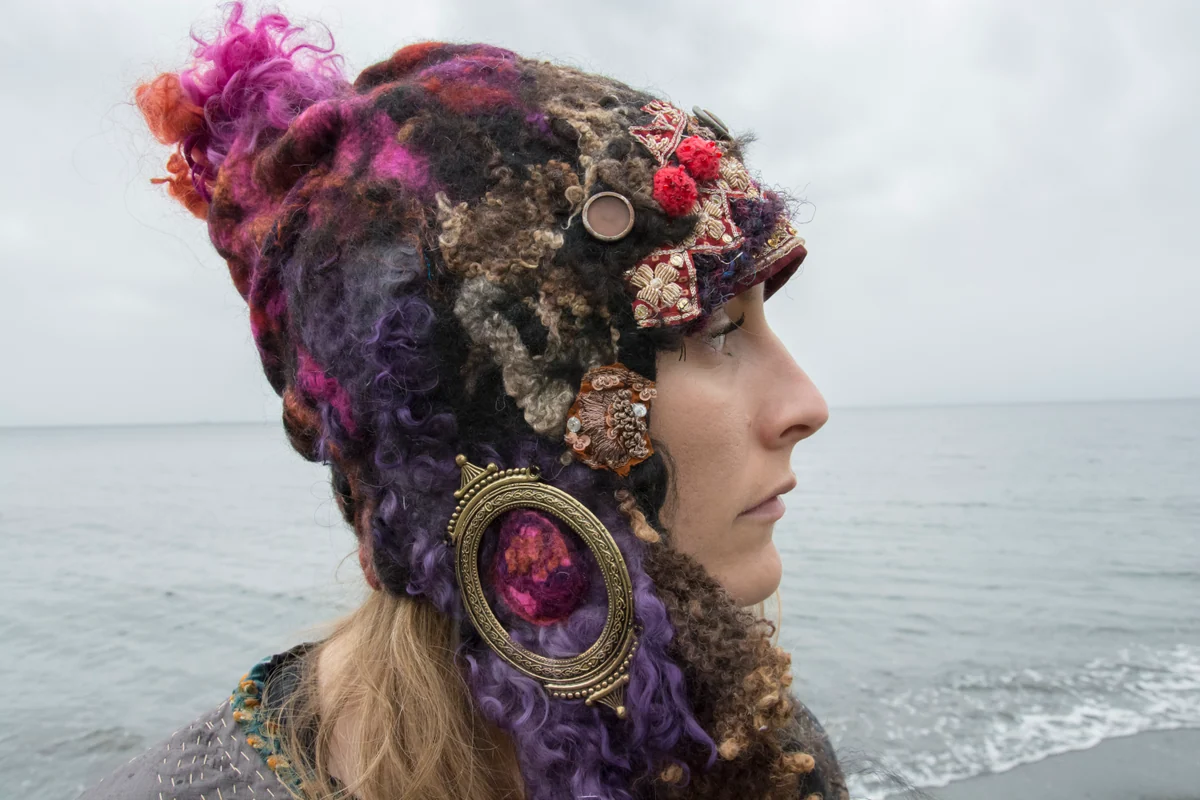 Model wearing dark textured fibre headwear with pink felt, patterned cloth and circular embellishments.
