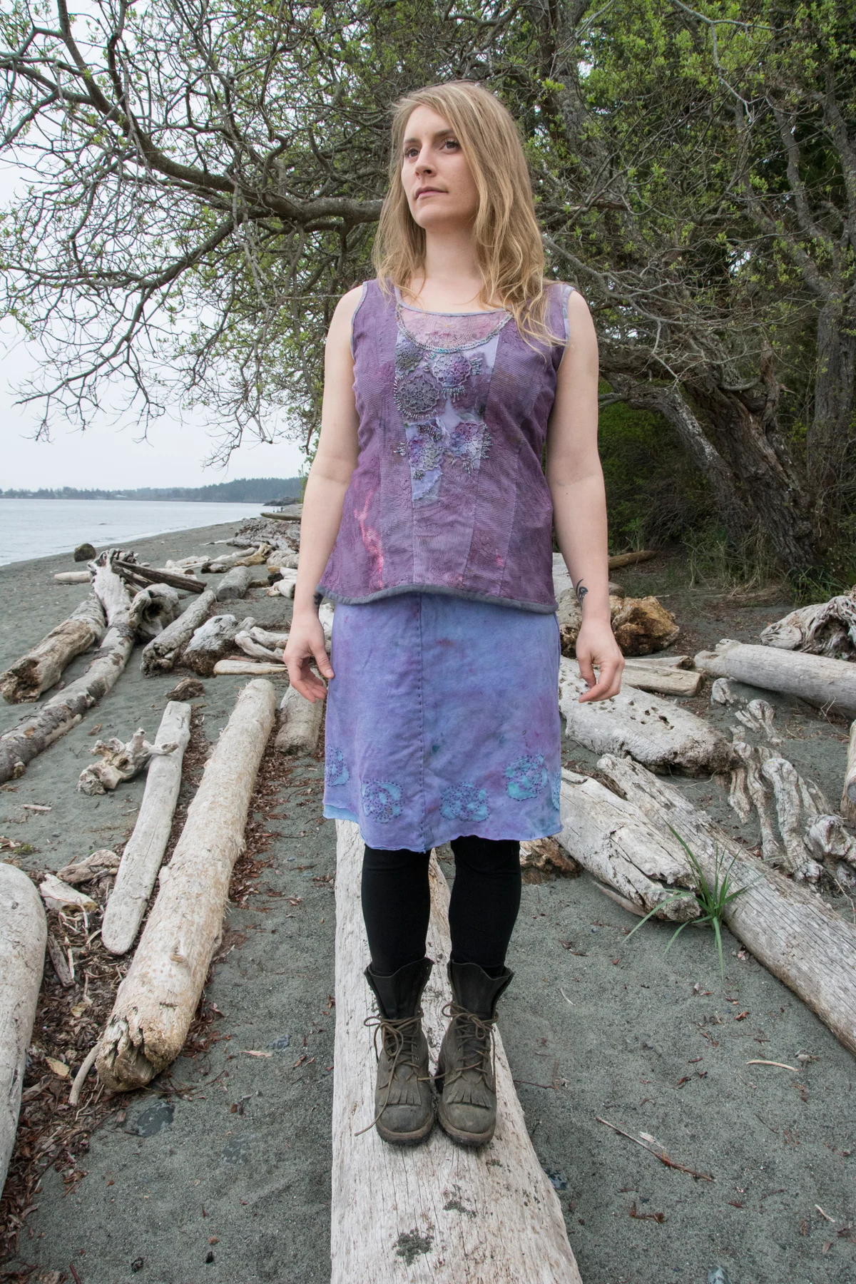 Model wearing a purple sleeveless textile top and skirt on a driftwood beach.
